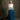 A woman stands in a studio wearing a loose white blouse with rolled sleeves and a long teal skirt. She is posed against a textured dark gray-brown backdrop, with a small dark box at the lower left.