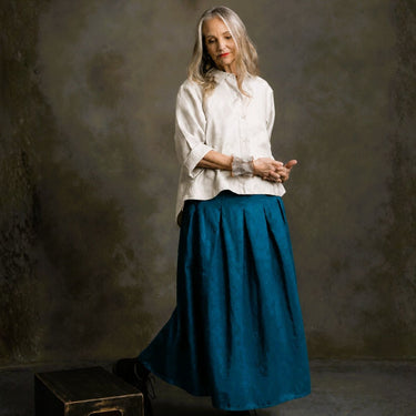 A woman stands in a studio wearing a loose white blouse with rolled sleeves and a long teal skirt. She is posed against a textured dark gray-brown backdrop, with a small dark box at the lower left.