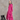 An older woman with gray hair stands in a studio wearing a bright pink long coat over a white dress, with a small black stool at her side against a textured gray backdrop.