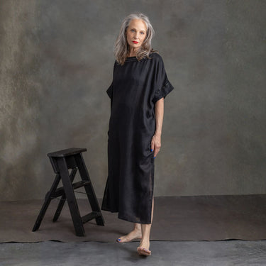A woman stands barefoot in a studio wearing a loose black satin-like dress or robe with short sleeves and side slits. A small black stool sits beside her against a textured gray backdrop.