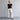 An older woman stands barefoot against a light gray wall, wearing a black short-sleeve top and wide-leg off-white pants.