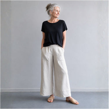An older woman stands barefoot against a light gray wall, wearing a black short-sleeve top and wide-leg off-white pants.