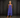 A woman wearing a sleeveless, tiered blue maxi dress with side pockets, standing against a dark textured studio backdrop.