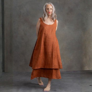 A woman with gray hair and red lipstick wearing a rust-orange linen dress, standing barefoot against a textured gray studio wall.
