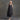 An older woman with gray hair stands barefoot against a textured gray studio backdrop, wearing a loose black sleeveless tunic dress with matching black pants.
