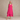 A woman with gray hair wearing a sleeveless, tiered magenta dress, standing barefoot against a textured beige studio backdrop.