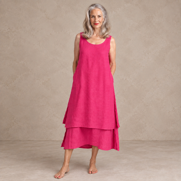 A woman with gray hair wearing a sleeveless, tiered magenta dress, standing barefoot against a textured beige studio backdrop.