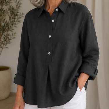 A woman wearing a dark charcoal gray linen button-up shirt with white buttons and white pants, standing in a softly lit indoor setting.