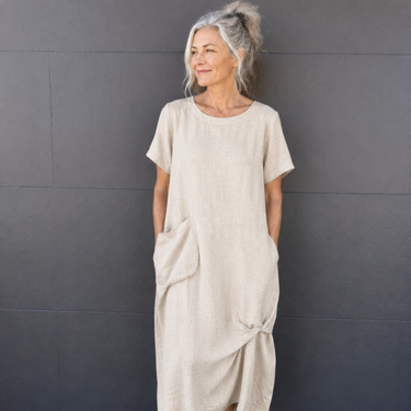 A woman with gray hair tied up stands against a dark gray paneled wall wearing a loose, short-sleeve linen midi dress in a light beige color. The dress has side pockets and a gathered, asymmetrical hem detail.