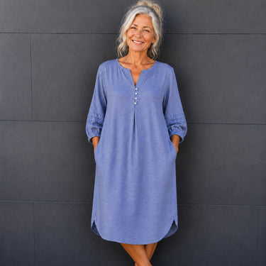A smiling older woman stands against a dark gray paneled wall wearing a loose, long-sleeve midi dress in a dusty periwinkle blue color. The dress features a split V-neckline with decorative buttons, gathered cuffs, and side pockets.